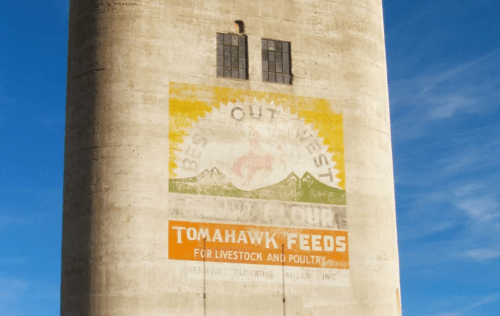 A weathered silo with faded advertisements for "Tomahawk Feeds" and "Best Out West" against a blue sky.