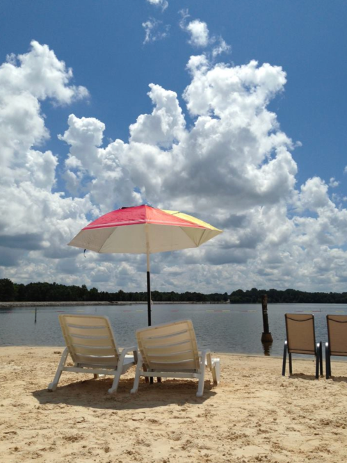Two lounge chairs under a colorful umbrella on a sandy beach by a calm lake, with fluffy clouds in a blue sky.