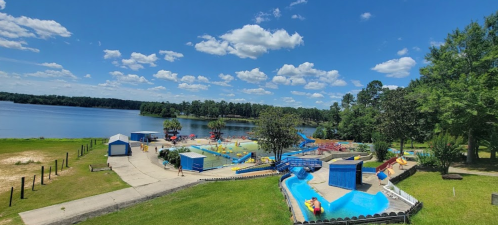 A sunny view of a water park by a lake, featuring slides, pools, and green trees under a blue sky with fluffy clouds.