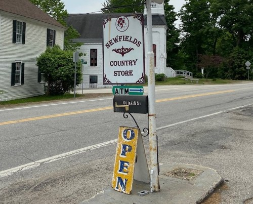 Sign for Newfields Country Store with "OPEN" displayed, located on a roadside near a church and trees.