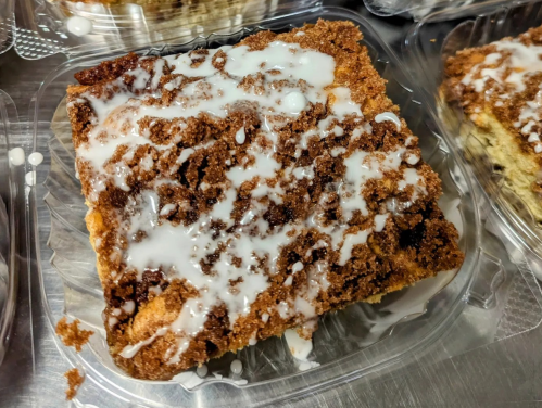 A square piece of coffee cake topped with a drizzle of white icing, placed in a clear plastic container.
