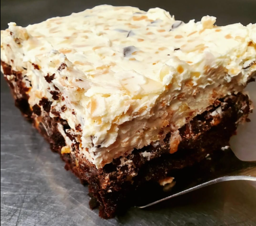A slice of layered dessert with chocolate base, creamy topping, and crunchy bits, served on a metal surface.