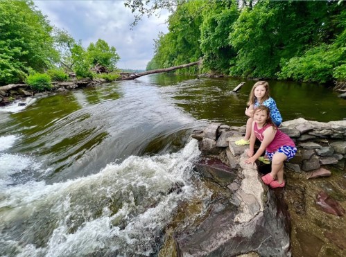 Two children sit on a rocky riverbank, surrounded by lush greenery and a flowing river under a cloudy sky.