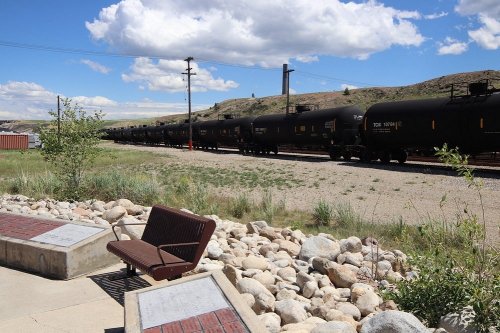 A long train of black tank cars passes by a bench and rocky landscape under a blue sky with clouds.