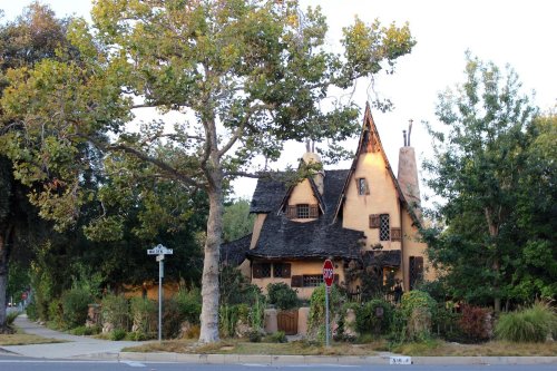 A charming, rustic house with a thatched roof, surrounded by lush greenery and trees, on a quiet street corner.