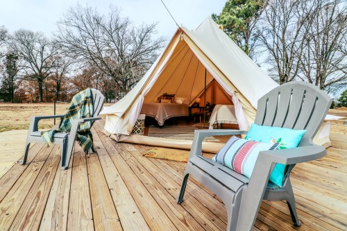 A cozy glamping tent on a wooden deck, with two chairs and a colorful pillow, surrounded by trees.