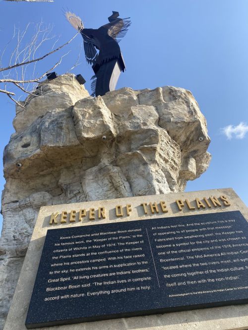 A tall statue of a figure with outstretched wings atop a rocky pedestal, with an informational plaque below.