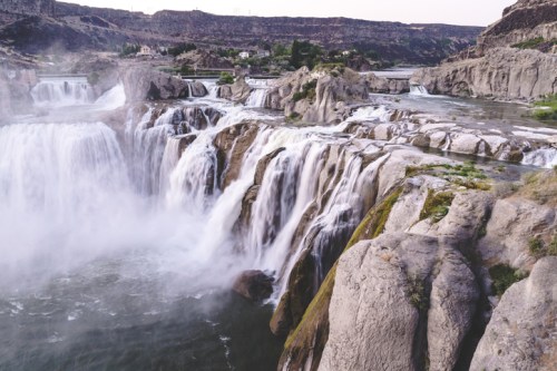 A stunning waterfall cascades over rocky cliffs into a river, surrounded by lush greenery and rugged terrain.