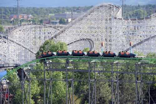 A roller coaster with riders ascending a steep track, surrounded by trees and a backdrop of other coaster structures.