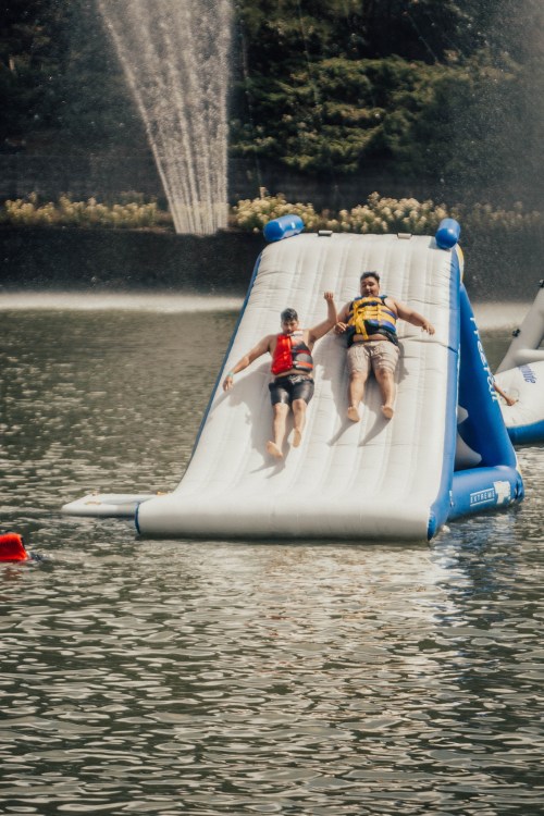 Two people slide down an inflatable water slide into a lake, surrounded by greenery and a fountain in the background.