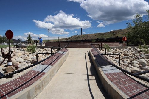 A pathway with brick accents leads through a rocky area under a blue sky with clouds, with people in the background.