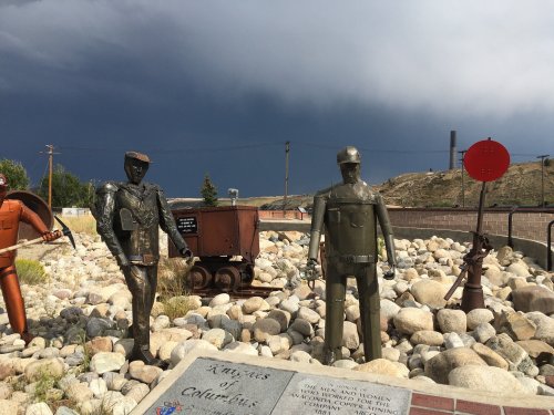 Metal sculptures of miners stand among rocks, with a dark sky and a red signal in the background.