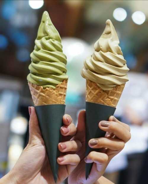 Two hands hold ice cream cones: one with green matcha swirl and the other with brown caramel swirl, both in dark cones.