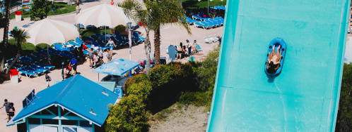 Aerial view of a water slide with a person sliding down, surrounded by palm trees and sun loungers.