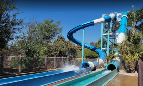 A colorful water slide with a twisting blue and white design, surrounded by greenery and a clear blue sky.