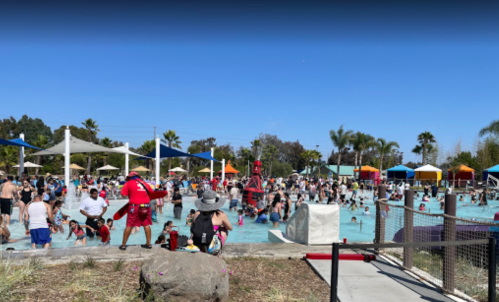 A crowded outdoor pool scene with people swimming, lounging, and enjoying the sun under clear blue skies.