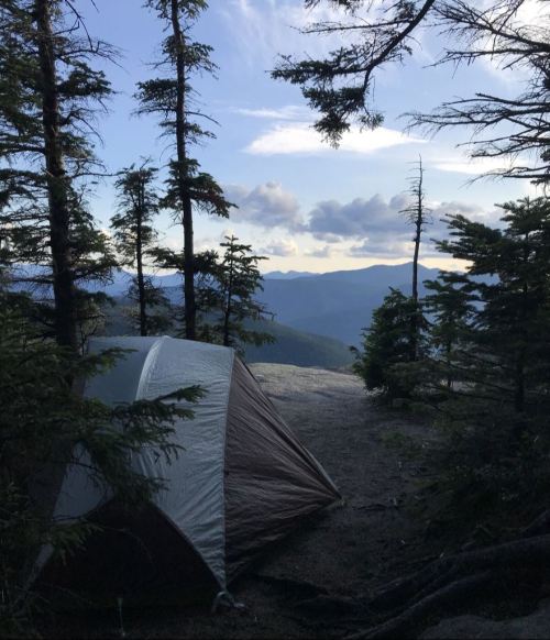 A tent set up in a forest clearing, surrounded by trees and mountains under a cloudy sky.