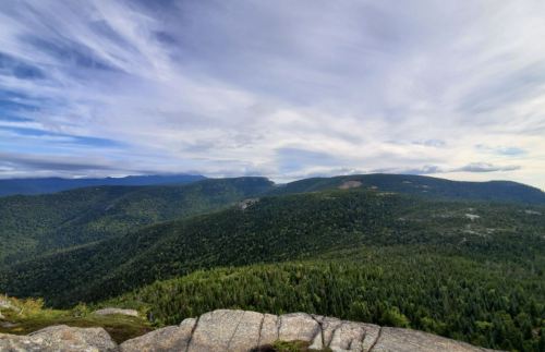 A panoramic view of lush green mountains under a cloudy sky, with rocky foreground and distant peaks.