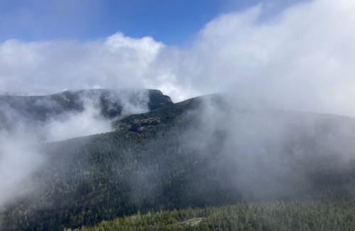 A misty landscape of mountains and dense forests under a blue sky with scattered clouds.