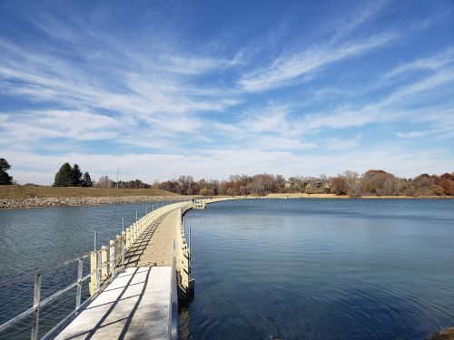 A wooden walkway extends over calm water, surrounded by trees and a clear blue sky with scattered clouds.