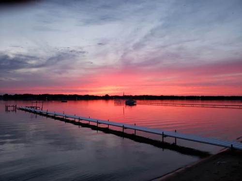 A serene sunset over a calm lake, with vibrant pink and purple hues reflecting on the water and a dock extending into the scene.