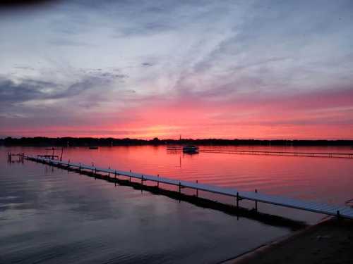 A serene sunset over a calm lake, with vibrant pink and purple hues reflecting on the water and a dock extending into the scene.