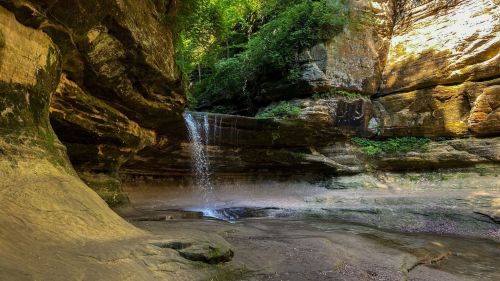 A serene waterfall cascades into a rocky canyon surrounded by lush greenery and sunlight filtering through the trees.