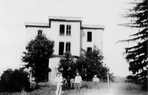 Black and white photo of a large, old building with four people standing in front, surrounded by trees and grass.