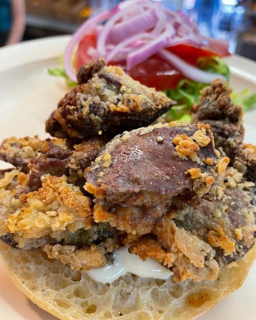 A plate featuring crispy fried meat on a bun, topped with mayonnaise and garnished with lettuce, tomato, and red onion.