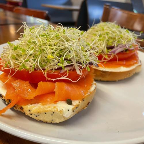 A bagel topped with cream cheese, smoked salmon, tomato slices, and a generous layer of sprouts on a white plate.