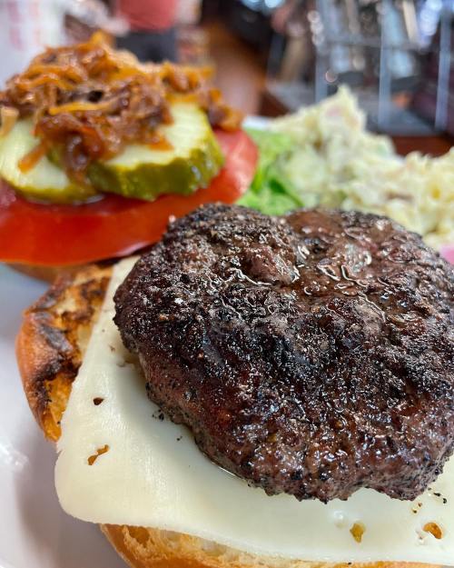 Close-up of a burger with a grilled beef patty, melted cheese, pickles, and a side of coleslaw on a plate.