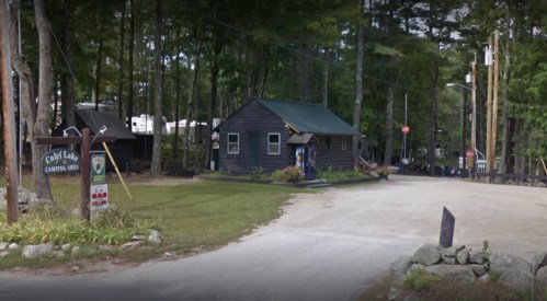 A rustic cabin at a campground, surrounded by trees, with a gravel road and sign indicating the area.