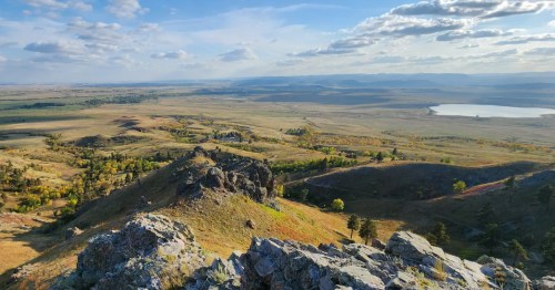 A vast, high-angle view over rolling, grassy hills and plains, with a distant lake and mountains under a blue, cloudy sky.