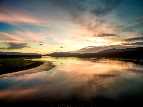 A serene landscape featuring a river at sunset, with colorful clouds reflecting on the water and mountains in the background.