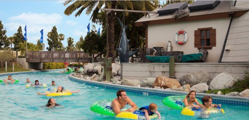 A lazy river with people floating on colorful tubes, surrounded by palm trees and a building in the background.