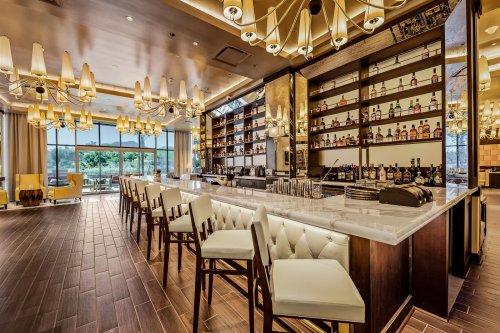 A modern bar with a sleek design, featuring white stools, a well-stocked shelf of bottles, and elegant lighting.