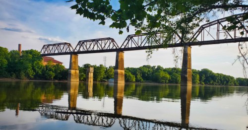 A rusted railway bridge spans a calm river, surrounded by lush greenery and a clear blue sky.