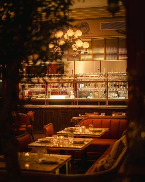 Cozy restaurant interior with warm lighting, elegant tables, and a bar lined with glasses in the background.