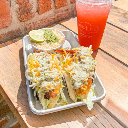 Two loaded tacos with toppings, a side of rice, and a refreshing drink on a wooden table.