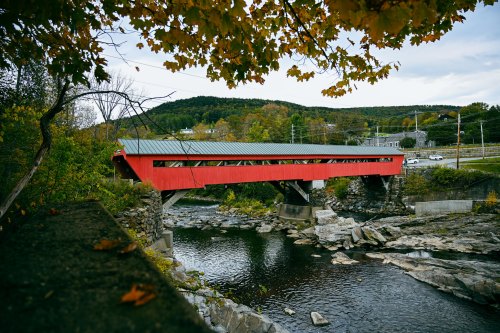 A red covered bridge spans a river, surrounded by autumn foliage and rolling hills in the background.