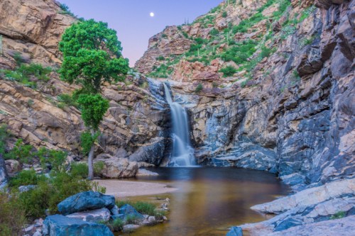 A serene waterfall cascades into a calm pool, surrounded by rocky cliffs and a lone tree under a twilight sky.