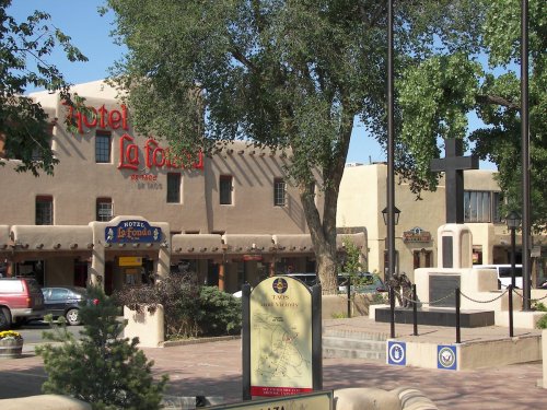 Historic plaza featuring Hotel La Fonda, a cross monument, and a map sign in a sunny, tree-lined area.