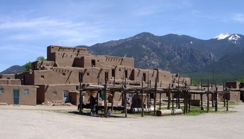 A traditional adobe pueblo with a mountainous backdrop under a clear blue sky.