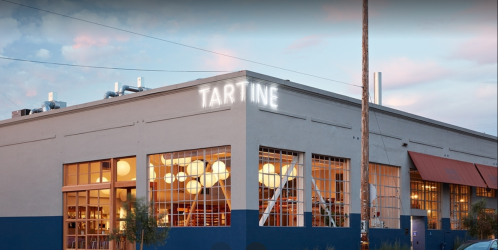 Exterior of Tartine bakery, featuring large windows and a neon sign, set against a colorful sky at dusk.