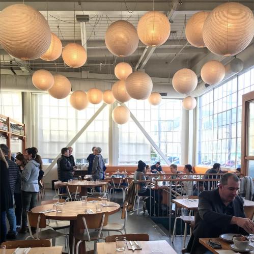 A bright restaurant interior with hanging lanterns, wooden tables, and patrons enjoying their meals and conversations.