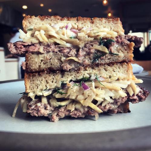 A thick sandwich with layers of beef patty, shredded vegetables, and sauce, served on toasted bread.