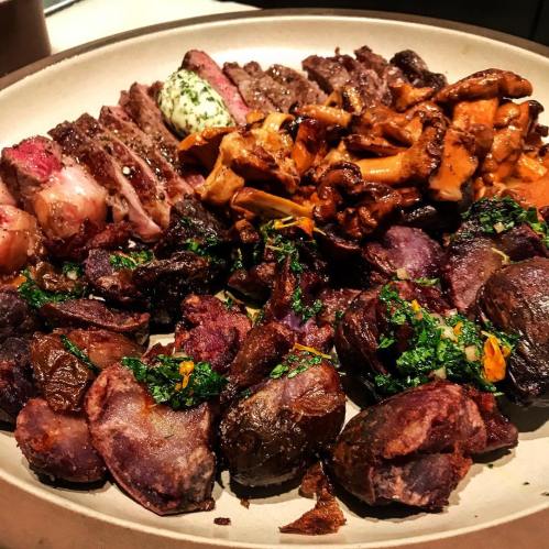 A plate of grilled meats, including steak and mushrooms, served with colorful roasted potatoes and herbs.