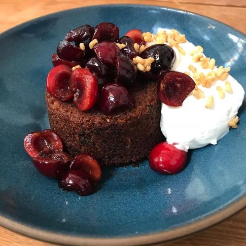 A round chocolate cake topped with cherries and served with a dollop of whipped cream on a blue plate.