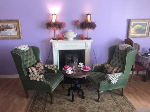 A cozy room with two green armchairs, a small table, and a fireplace, decorated with pink flowers and paintings.