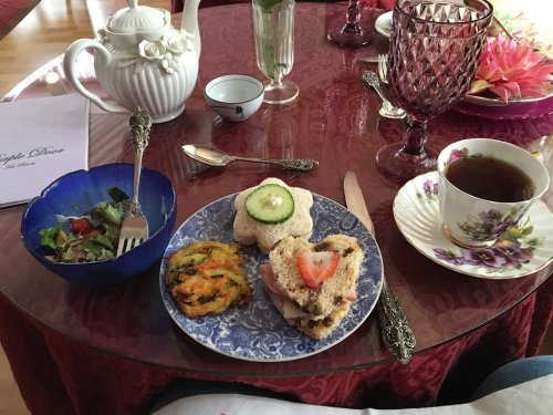A table setting with a teapot, cup of tea, sandwich, salad, and a colorful dessert on a decorative plate.